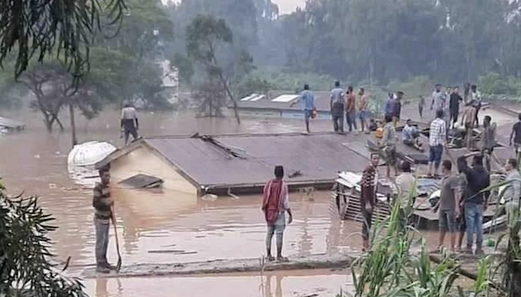 2 people stand on a floating log in the foreground while water reaches the roof of a building. Several people are standing on the roof.