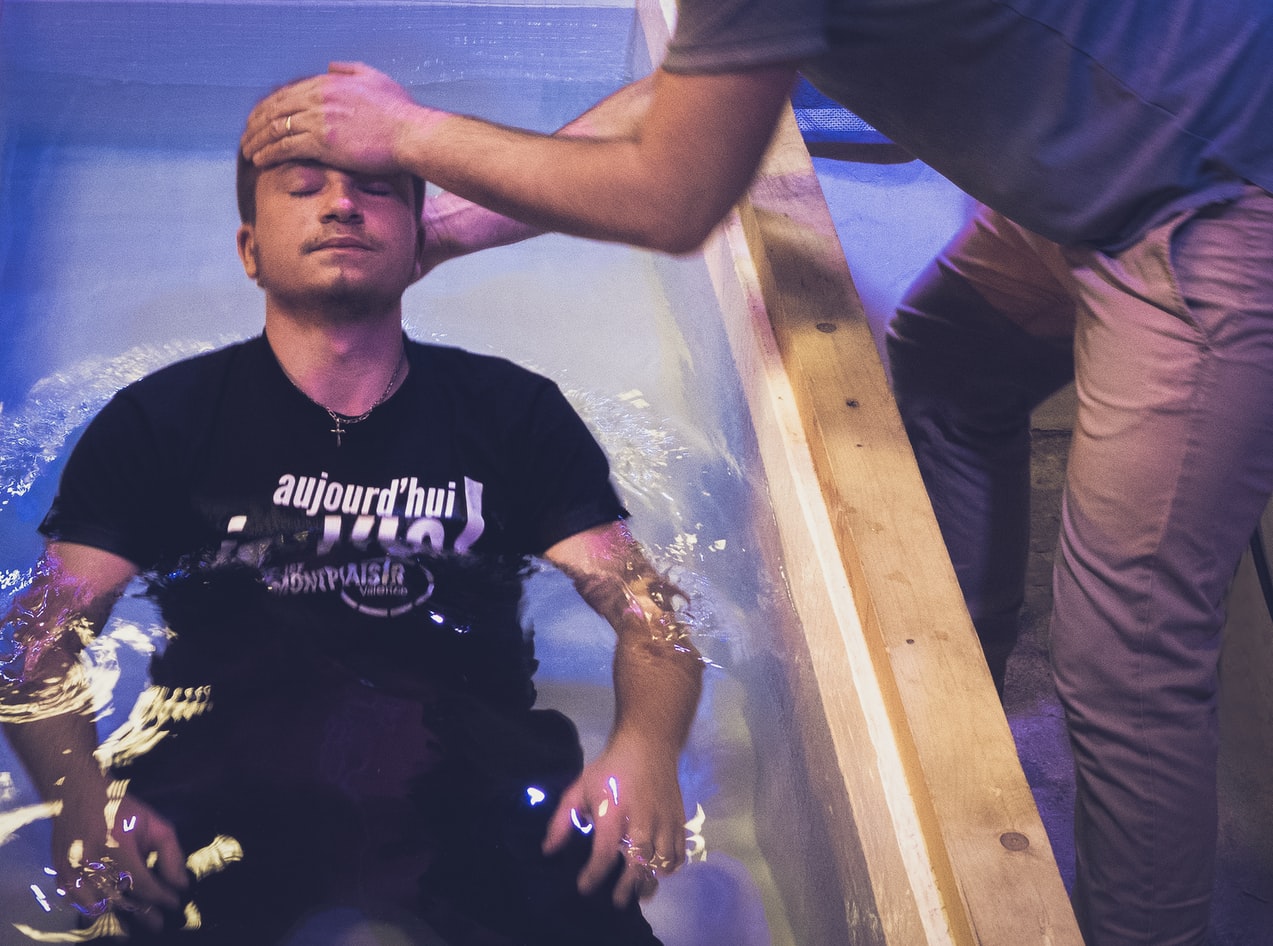 A pastor places his head on a man who is about to be baptized. The other man sits in a large tub of water with his head tilted back.
