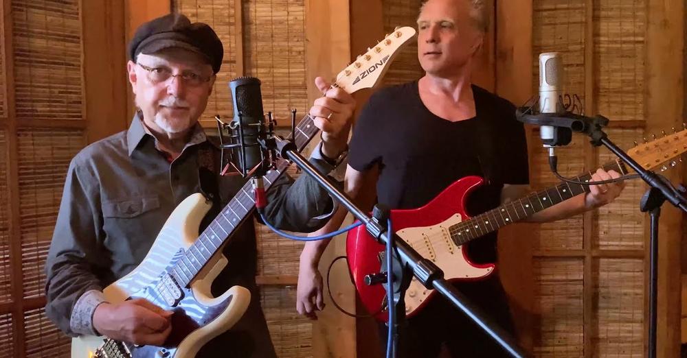 Two men hold electric guitars, and stand in front of microphones.