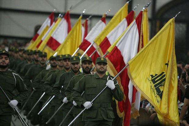 Soldiers dress in green uniforms and caps, march carrying yellow as well as white and red flags