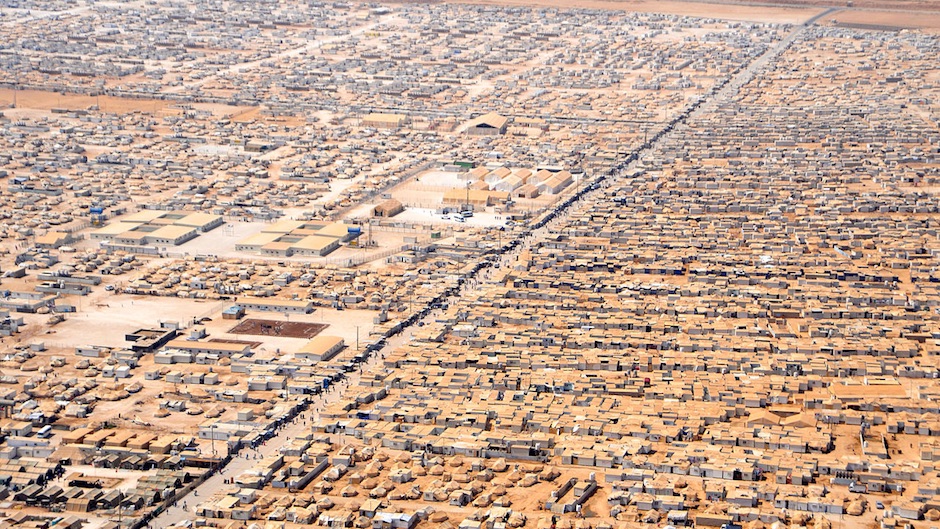 A refugee camp is seen from the air with many small buildings and tens with brown roofs.