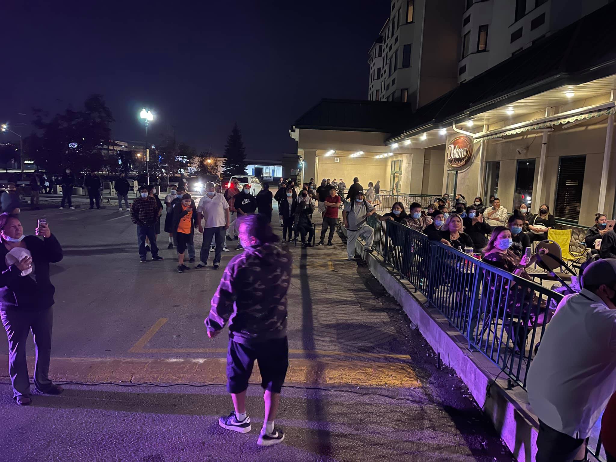 A group of people stand in a parking lot while others are seated inside a small area in front of the hotel