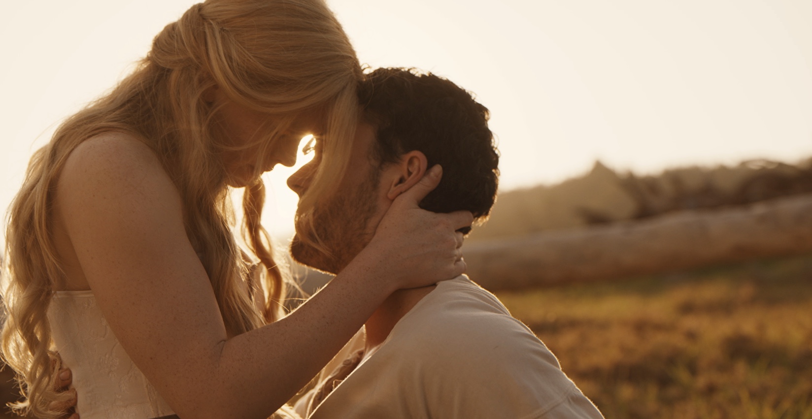 A man and a woman embrace with their foreheads touching and the sun shining behind them.