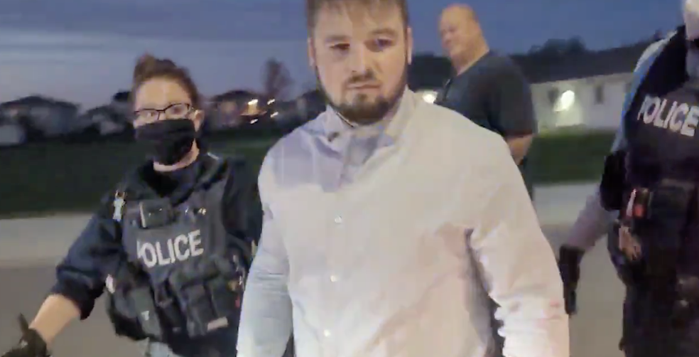 Tobias Tissen walks towards a police car, escorted by two officers in police vests and wearing masks