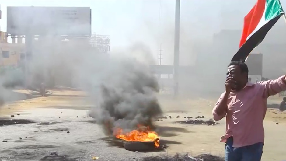 A man stands in the foreground holding a flag and covering his mouth with his hand while tires and other items burn in the street behind him and black smoke fills the air