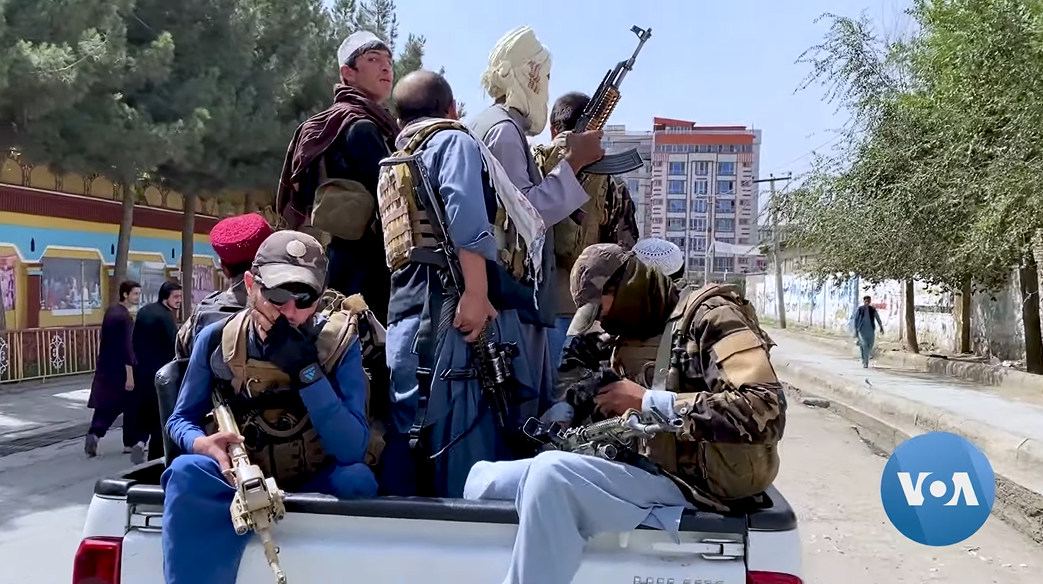 Several men in fatigues and holding guns sit in the box of a white pickup truck
