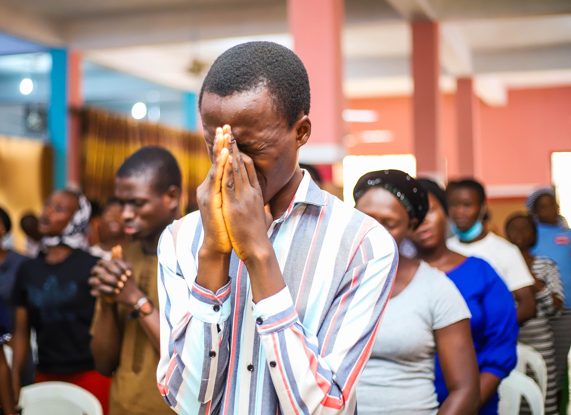 A group of people pray in a chruch, with a man in the forefront holding his hands together at his face.