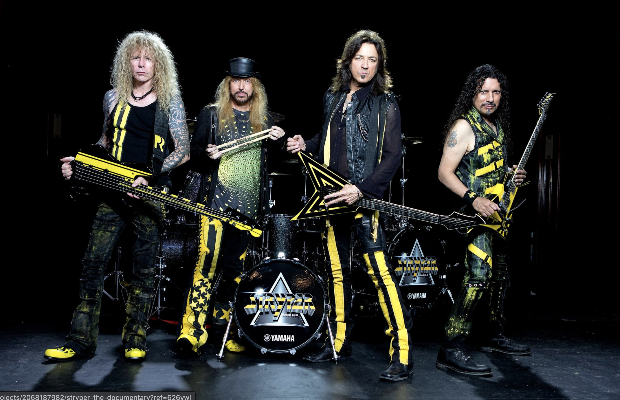 The 4 band members of Stryper standing with their instruments. All are dressed in black with some yellow stripes