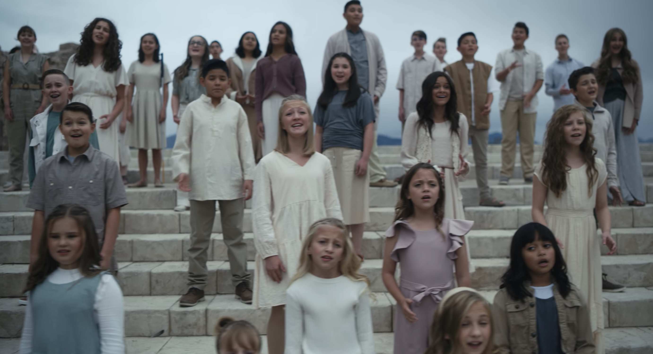 A number of young children stand on a large outdoor staircase and sing