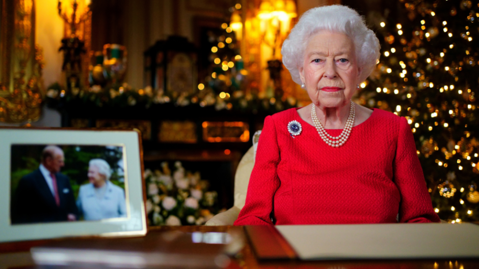 Queen Elizabeth sits in front of a Christmas tree in a red dress. A photo of her and her late husband sits slightly out of focus in the foreground