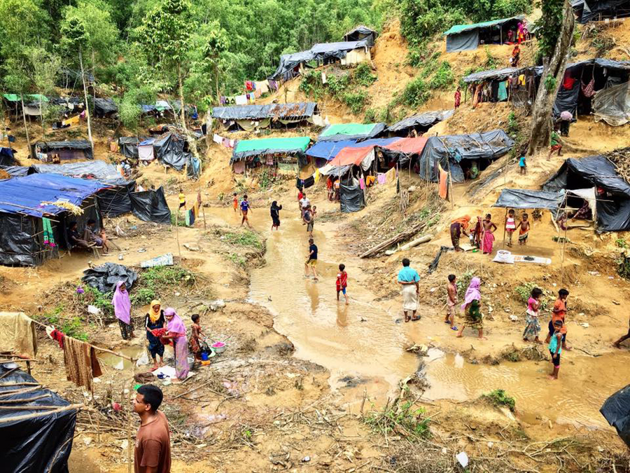 Shanties made out of sticks and tarps sit on the side of a dirt hill. People walk and stand around.