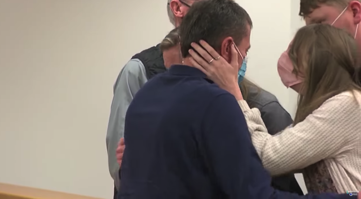 A young woman hold her hands to the face of a man while family looks on in a courtroom