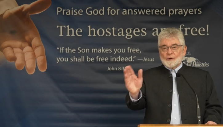 A man white grey hair and beard and wearing a black jackets speaks to reporters. He is lifting one hand in celebration and smiling
