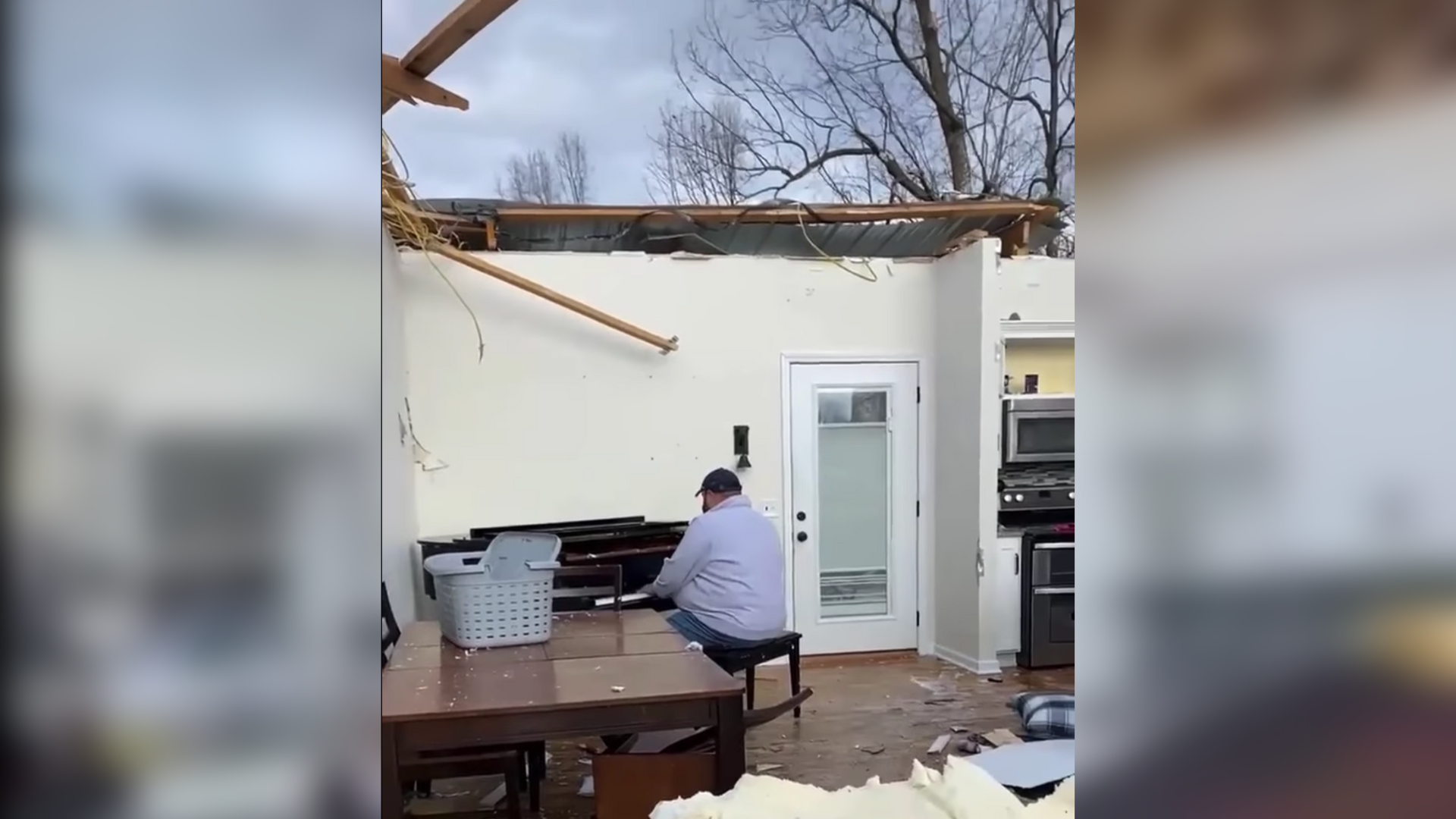 A man sits at a grand piano inside a home with the roof above him torn off. Damage from the home is on the ground around him.