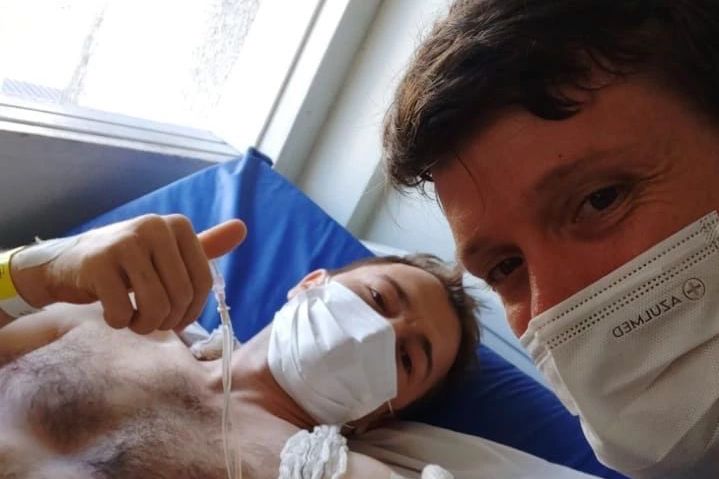 Arlen lays on a hospital bed while a friend looks into the camera - both of them give a thumbs up to the camera.