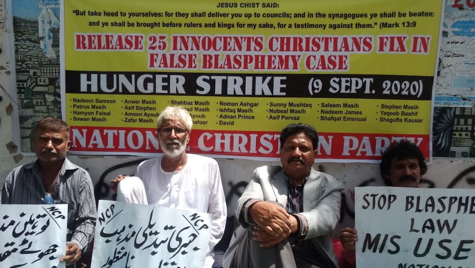 4 men sit on the ground in front of a large sign declaring a hunger strike and calling for the release of people accused of blasphemy