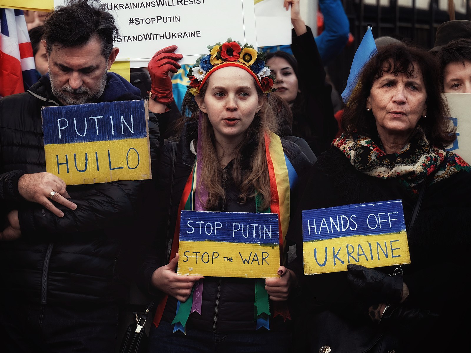 A man and 2 women hold blue and yellow signs with anti-war and anti-Putin messages