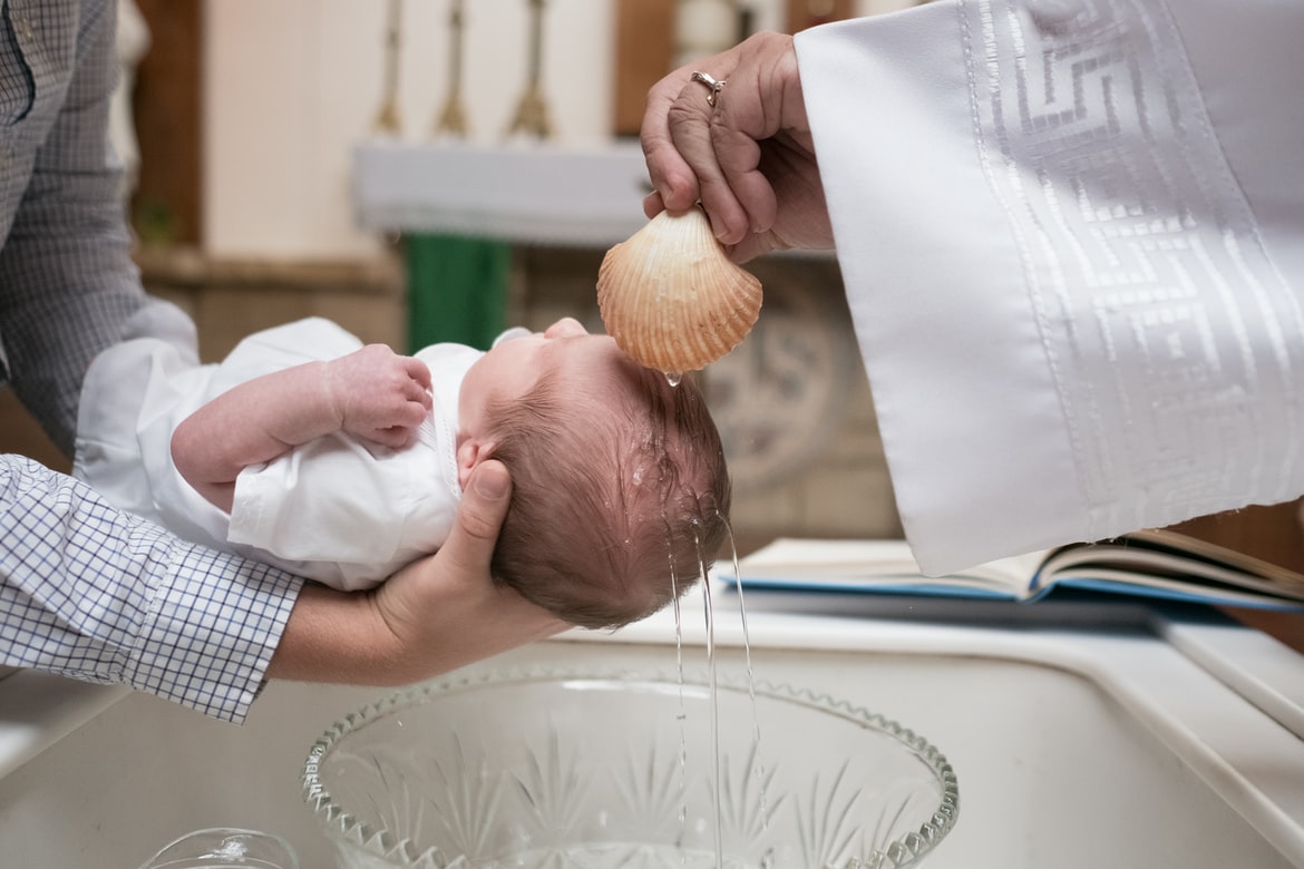 A priest pours water over the head of an infant above a baptismal font