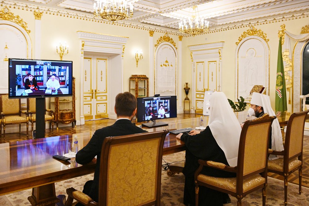 Russian Orthodox priests sit at a table facing screens with the Pope and a Cardinal talking to them. The Orthodox priests wear black robes and white kamilavka