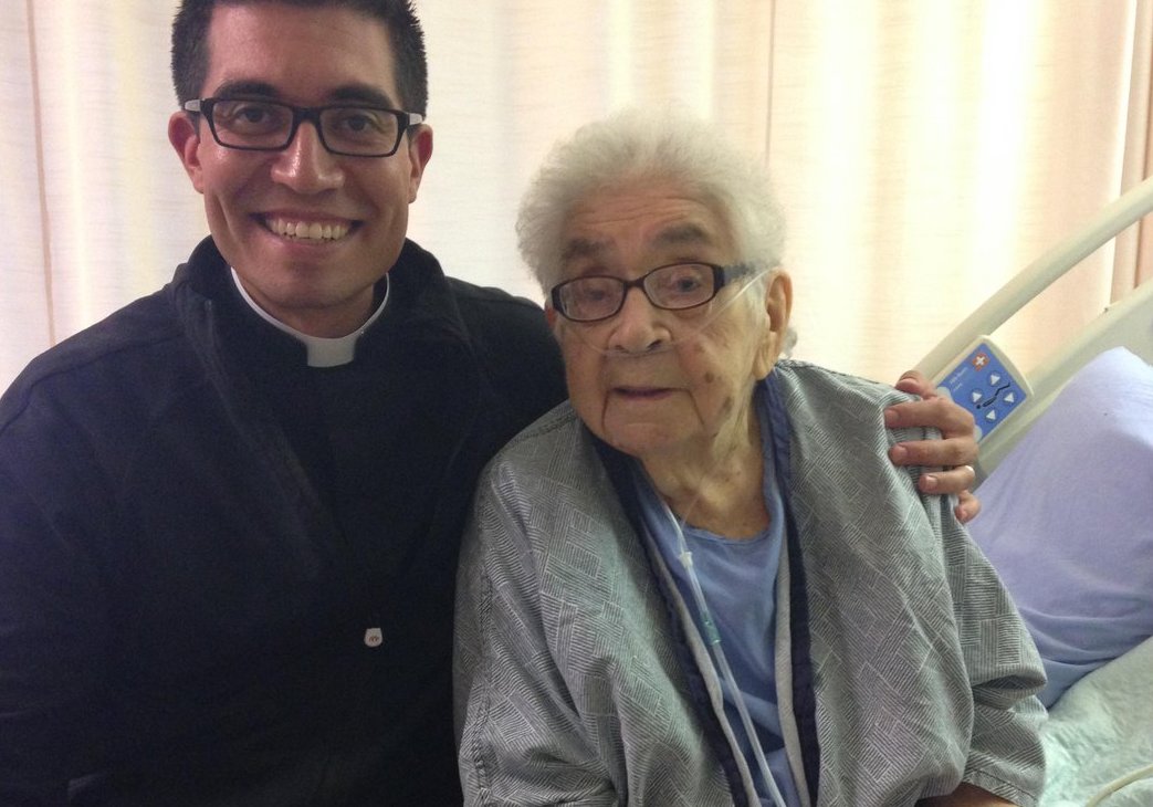 A man with medium skin tone, short black hair, and black glasses wears a clergy collar and has his arm around an elderly woman on a hosptial bed. She has medium skin tone and grey hair and glasses.