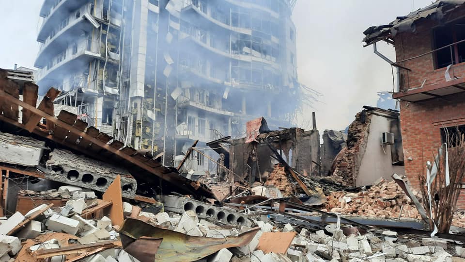 Rubble from a large building destoryed by an explosion. Smoke rises from the rubble and a damaged skyscraper sits in the background.