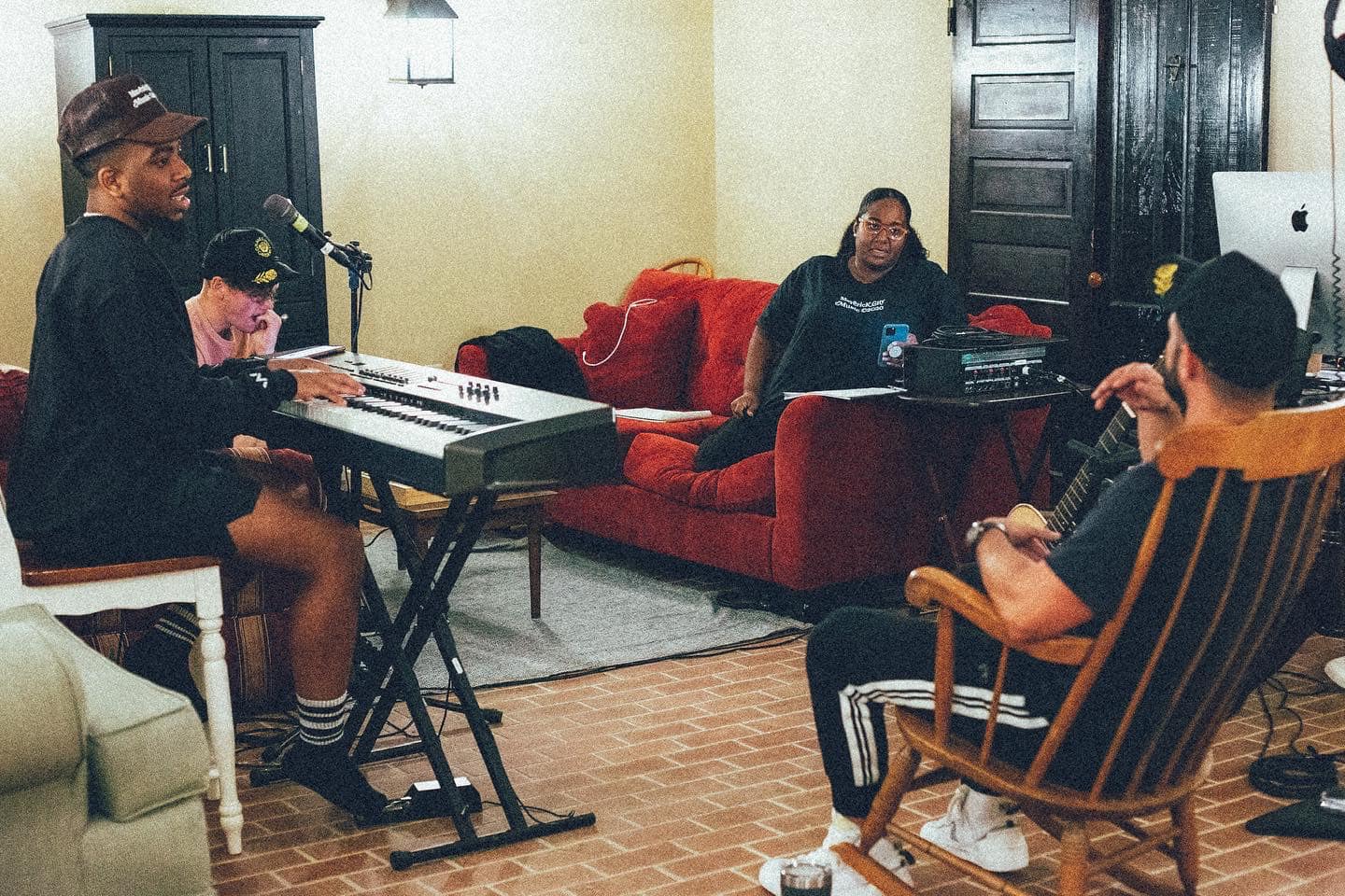 4 people sitting in a living room with instruments. Clockwise from the top, a woman with dark skin tone sits on a red couch , a man with light skin tone sits in an old wooden rocking chair, a man with dark skin tone sits on a wooden chair behind a keyboard, and a man with light skin tone holds his left hand under his chin.