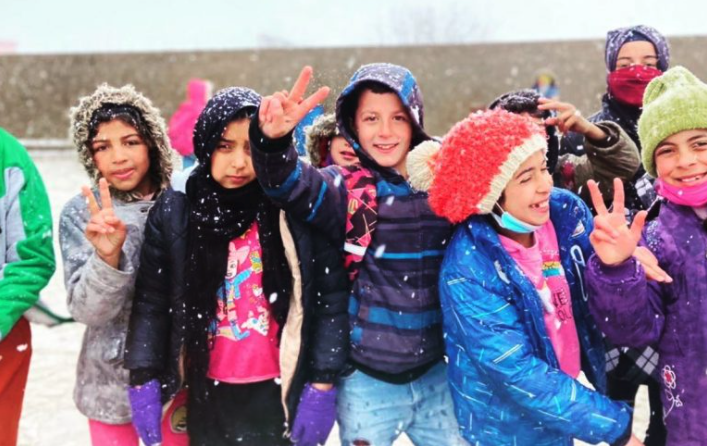A group of kids hold up peace signs and smile, dressed in bright clothes, as snow falls