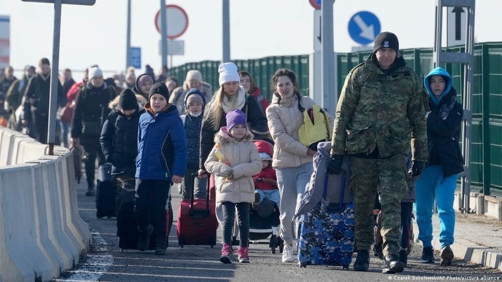 Large lines of people dressed in winter jackets walk down a sidewalk with most carrying or pulling suitcases.