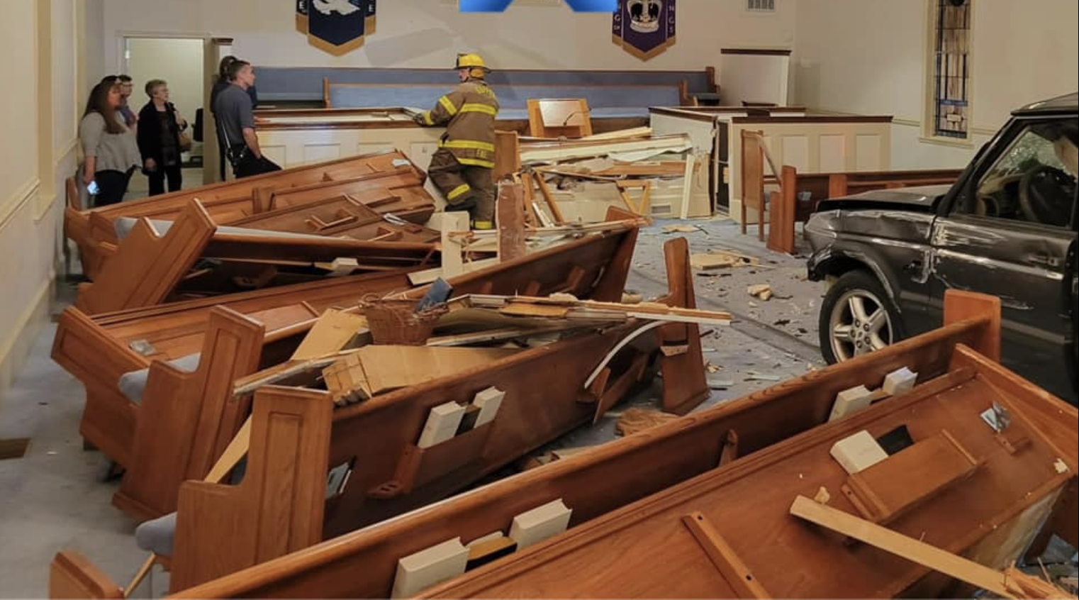 A car in the aisle of a church sanctuary. Broken pews are splinttered on the left side. A firefighter walks at the front of the sanctuary as a few other people look on near a back entry.