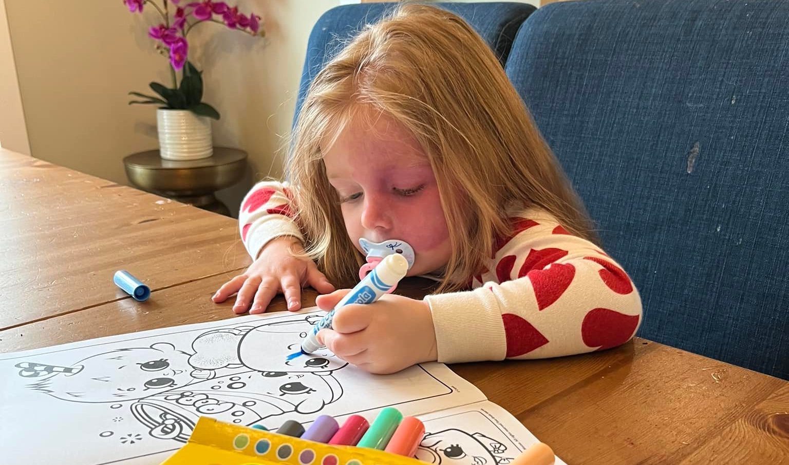 A three year old blonde haired girl with a large port-wine birthmark on her face. She has a pacifier in her mouth while she colours with markers at a kitchen table.