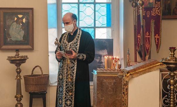 A priest in a black robe in an , Orthodox church sanctuary. Several small candles burn to his left, a blue stained glass window is behind him, and an icon painting of Jesus is on the wall to his right.