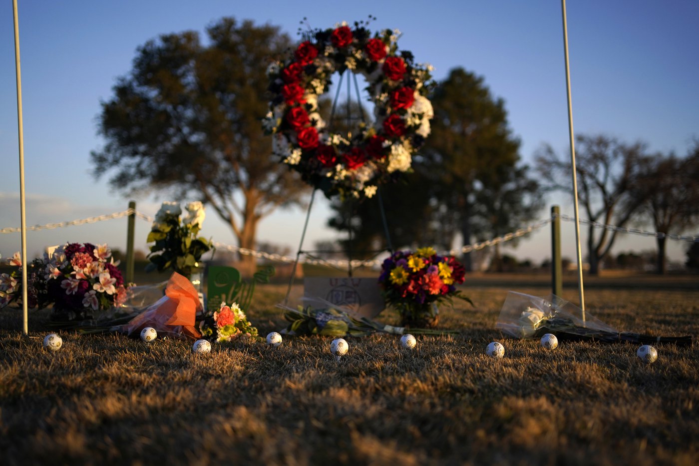 A makeshift memorial on the ground. A wreath is on a stand and golfballs and flowers are on the ground.