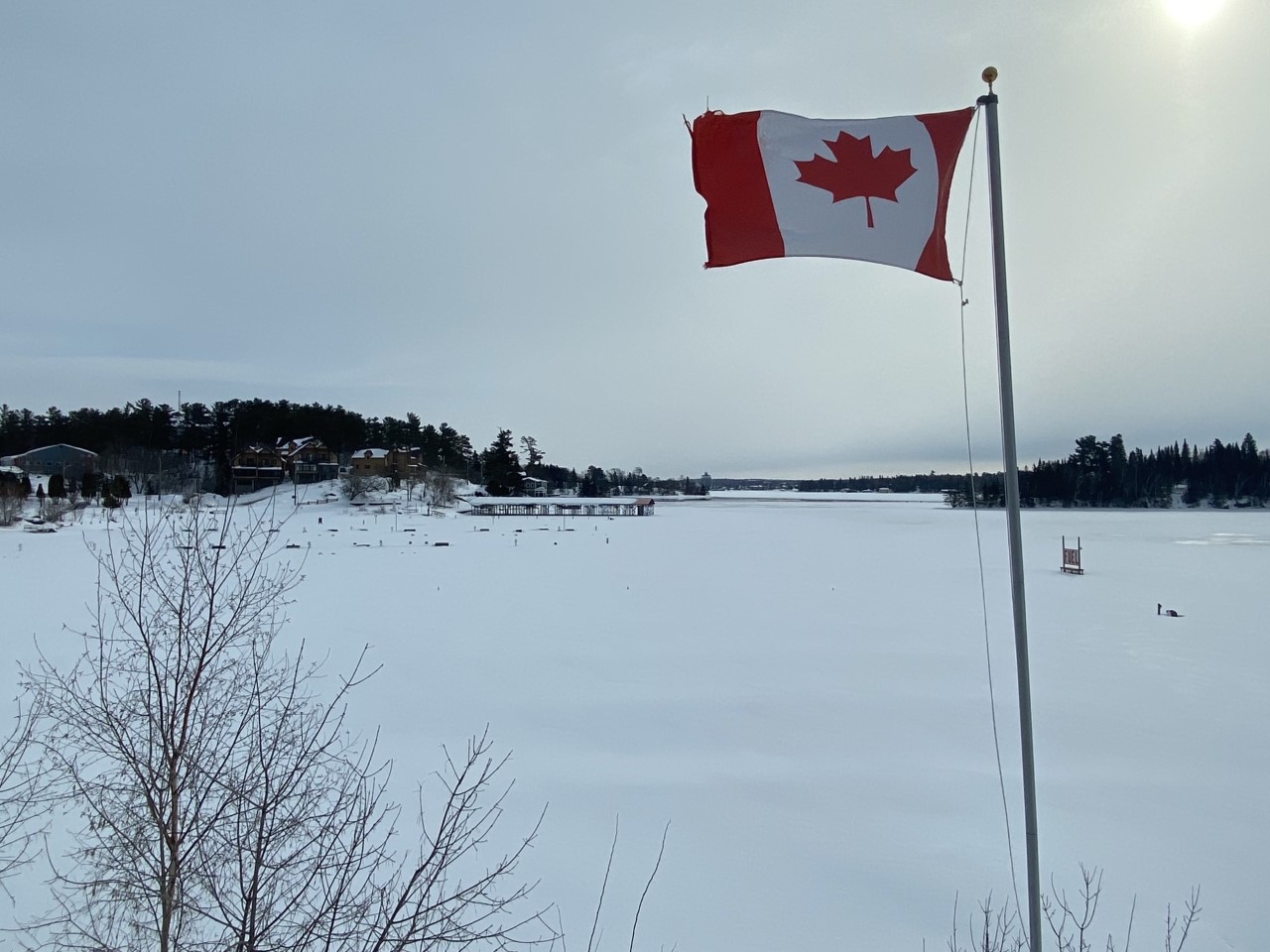 Picture of a frozen lake and light sun coverage, a Canadian flag is also in the foreground. 