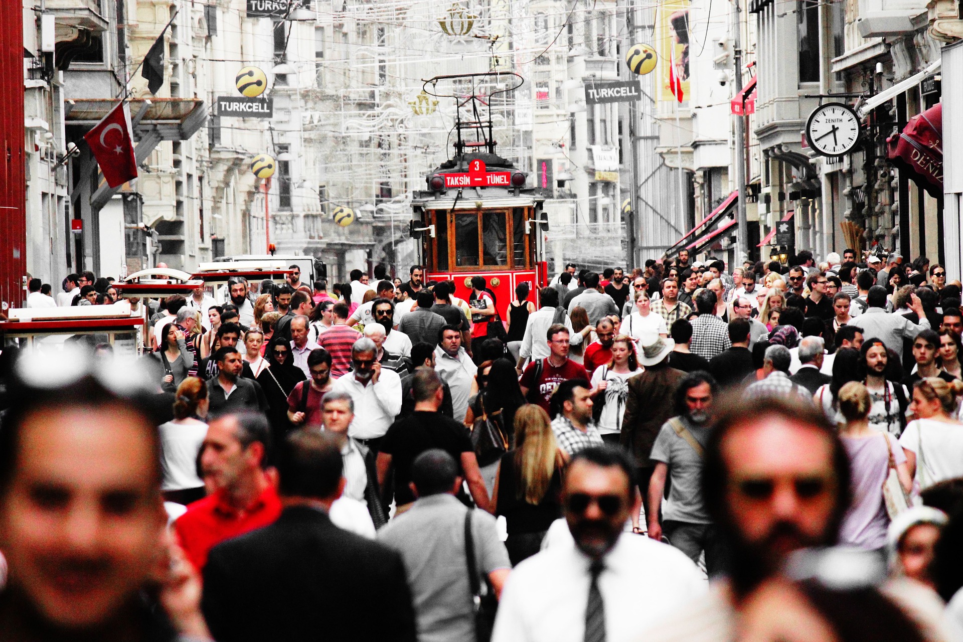 Large crowds walk on a street between tall older buildings. There is a red streetcar in the background.