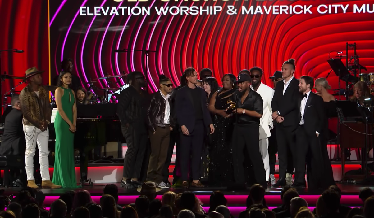 A large group of people stand on the stage at the GRAMMYs to accept their award