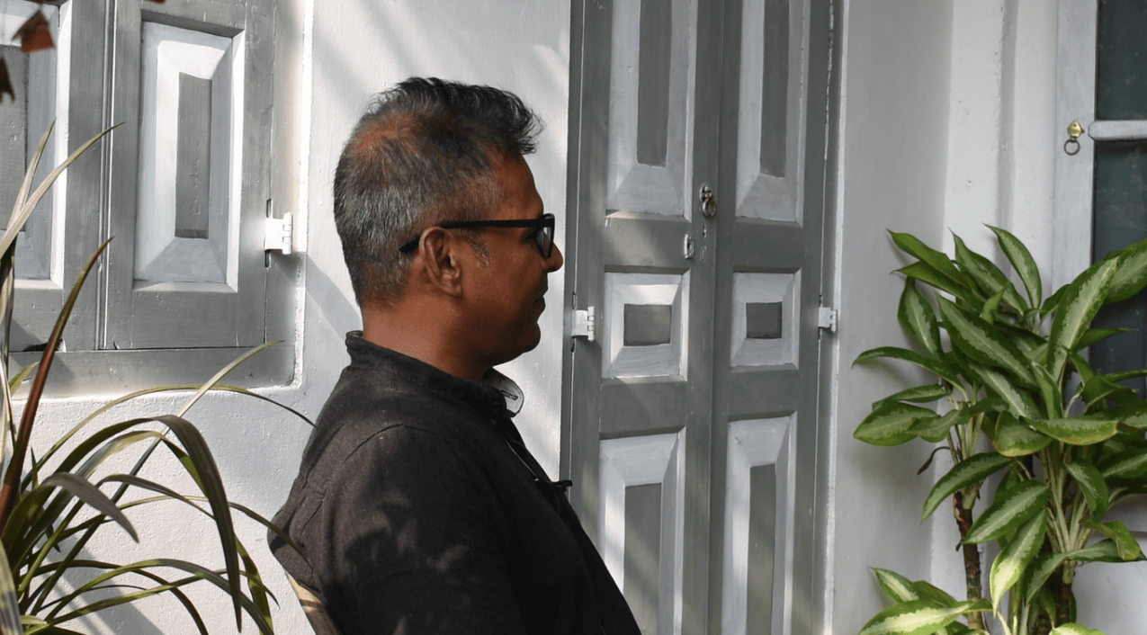 A side profile of a man with medium skin tone and short grey hair sitting on a chair on a porch in front of a white building with a grey door.