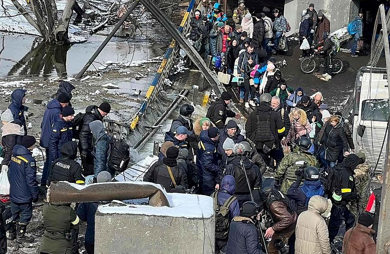 A line of people wait to get on buses amid destruction