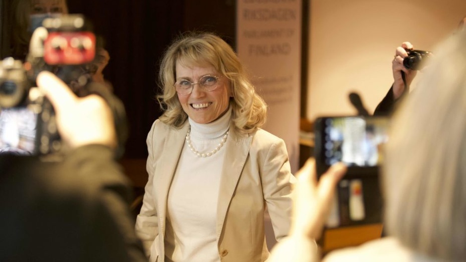 A Woman with light skin tone and blonde hair smiles while several cameras take her photo. She wears a beige blazer and white turtle neck.