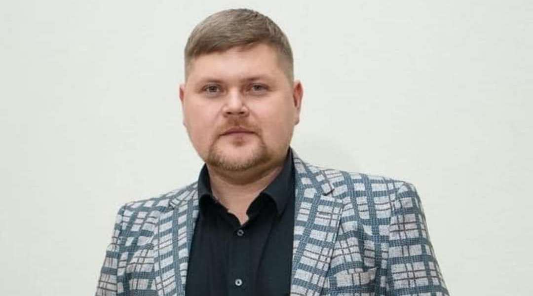 A man with light skin tone and light brown hair and goatee poses for a portraite. He wears a light blue sport coat with dark blue stripes and a black dress shirt.