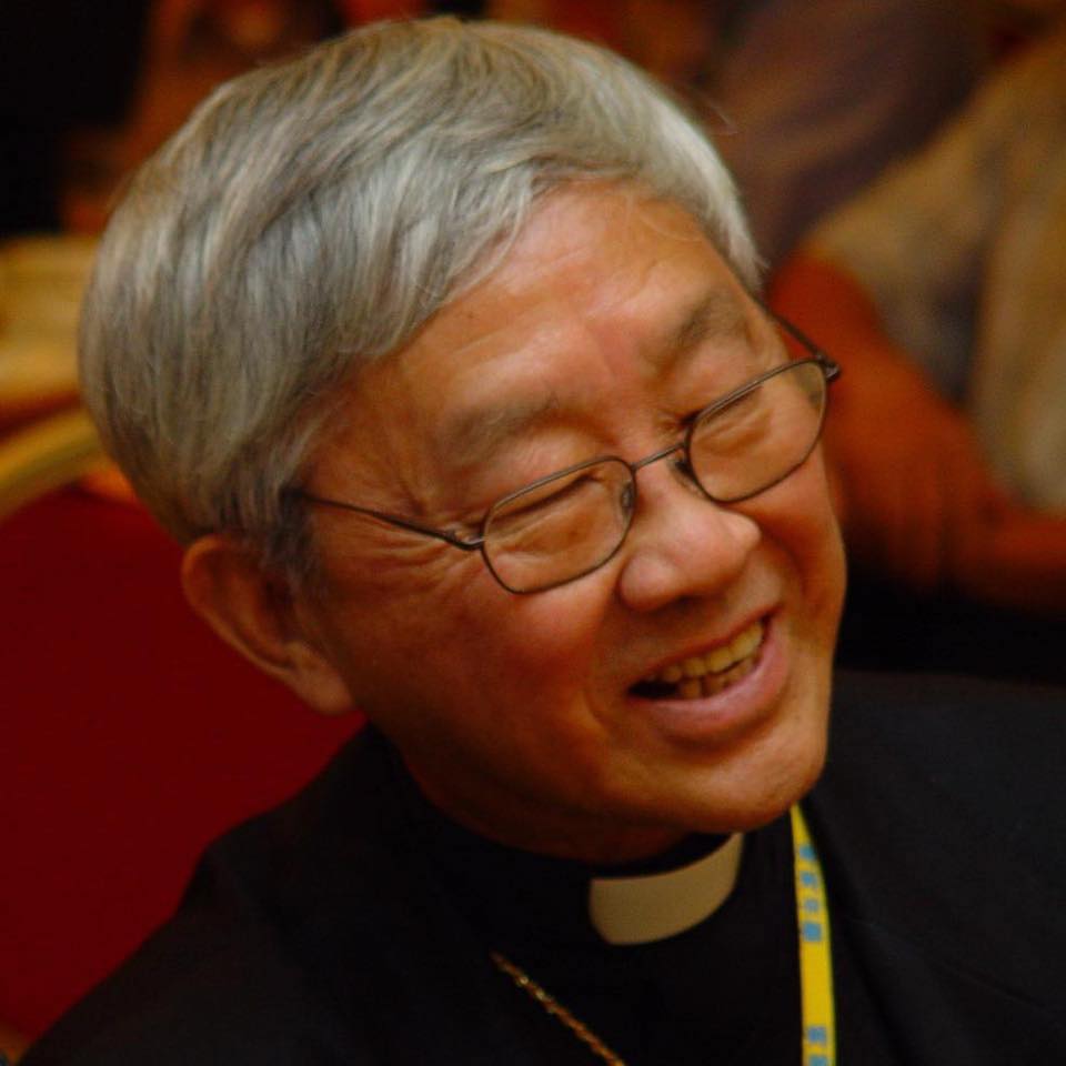 A man with medium skin tone and grey hair with glasses smiles. He is wearing a black shirt with a white clergy collar