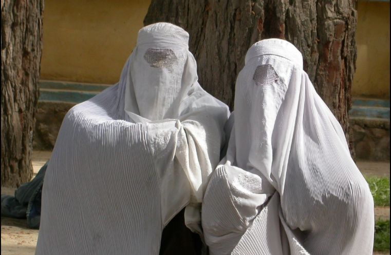 two figures in full length white Burkas, sitting on the side of a street