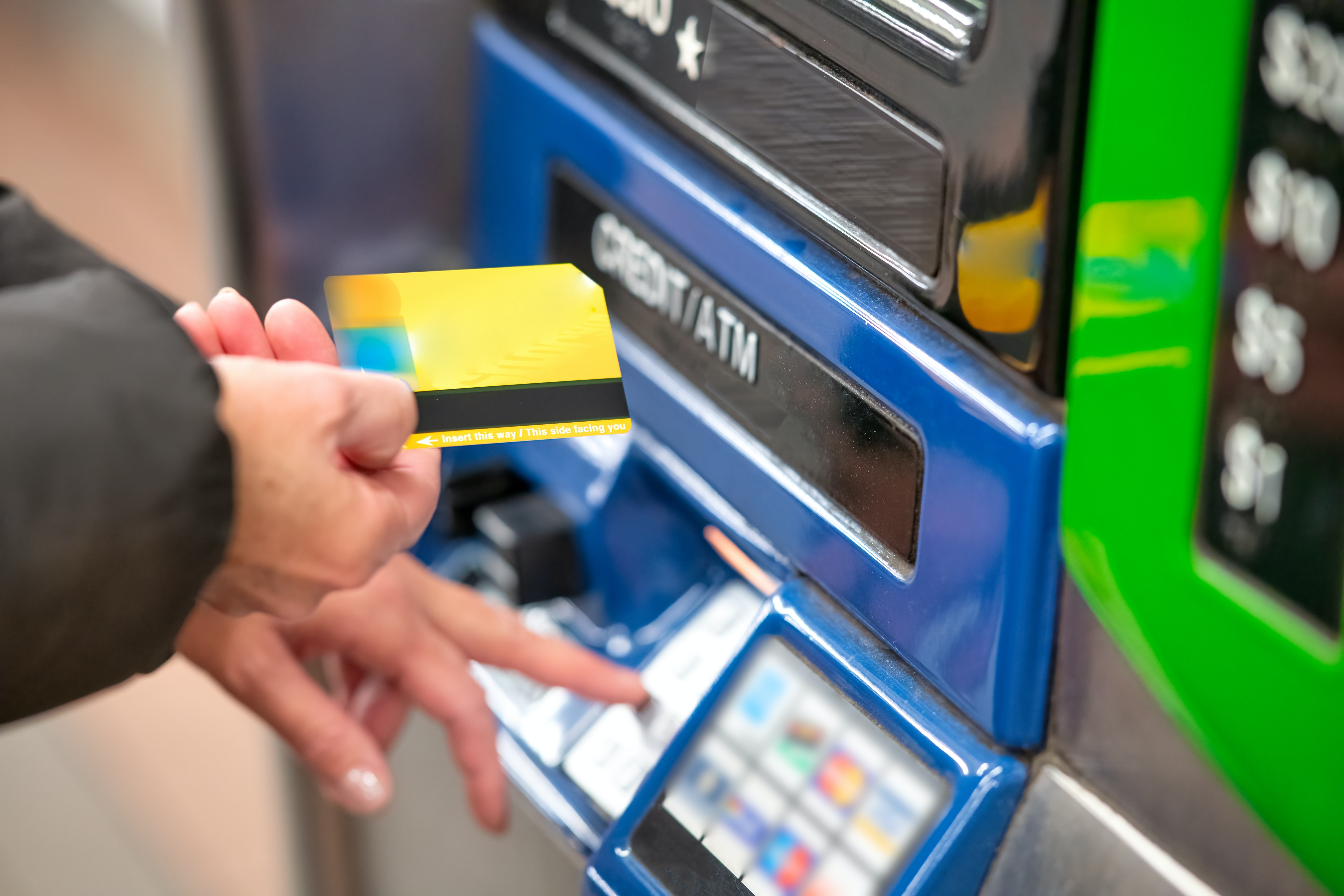 A hand is seen holding a generic yellow bank card while another hand punches numbers on an ATM keypad