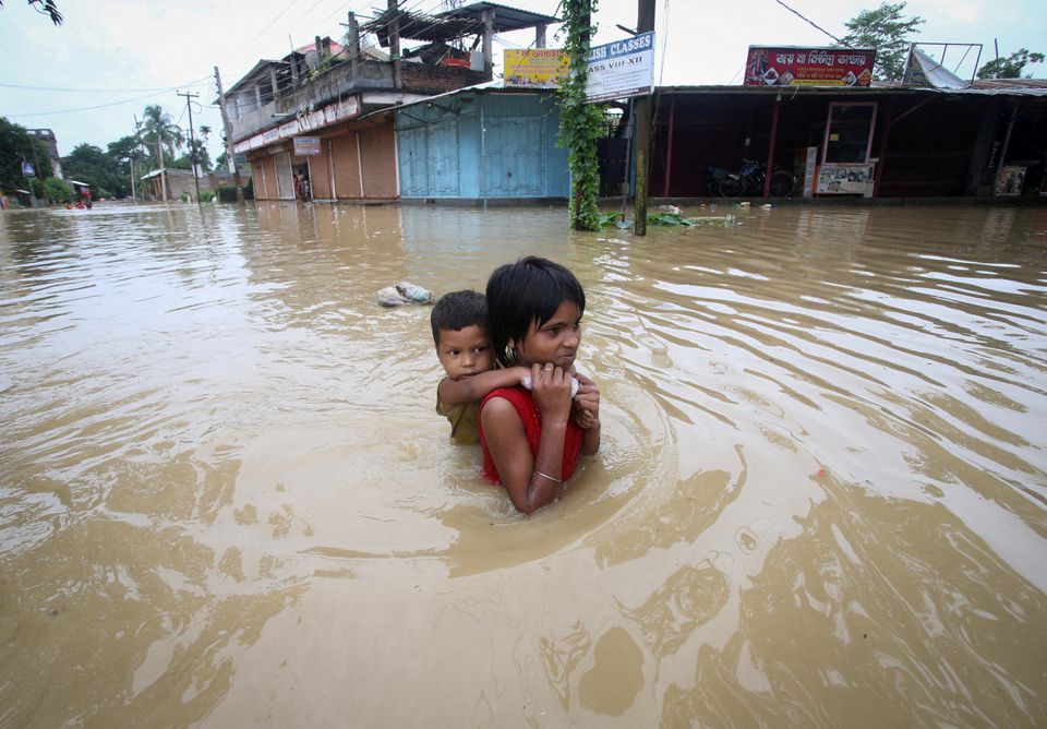 A small child about 10 to 12 years old, with medium skin tone, wades through flood waters in a street while carrying a pre-school aged child on their back