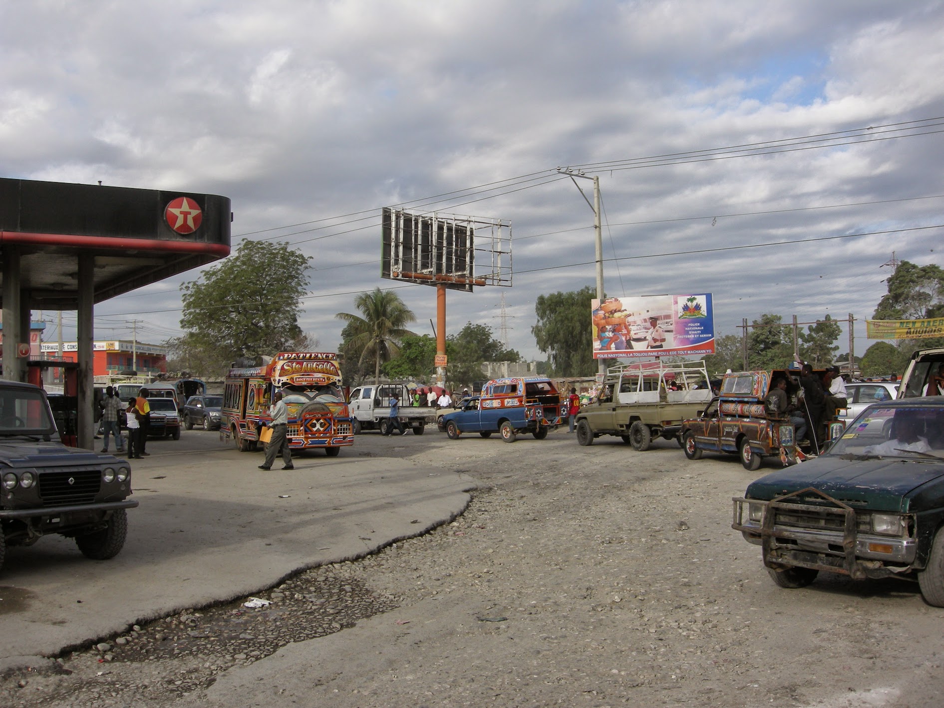 Old vehicles drive on a curve in the road. A Texaco gas station is on the left-hand side of the picture