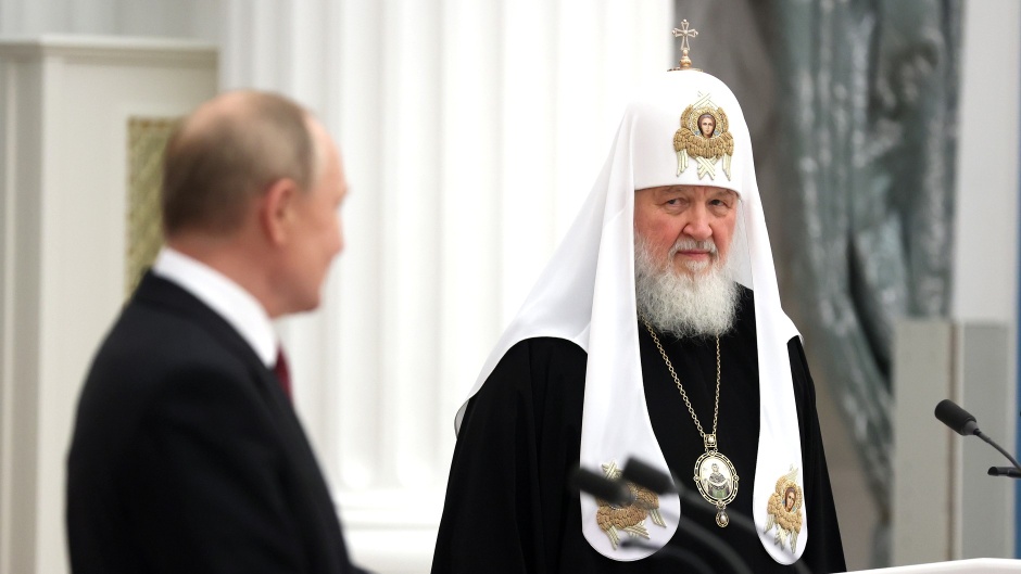 A 45 degree angle shot of two men standing in a room. The man on the lift has light skin tone and short balding hair - he wears a dark suit. The man on the left wears a black robe and a tall white hat with a white robe on top - he has a long grey beard.