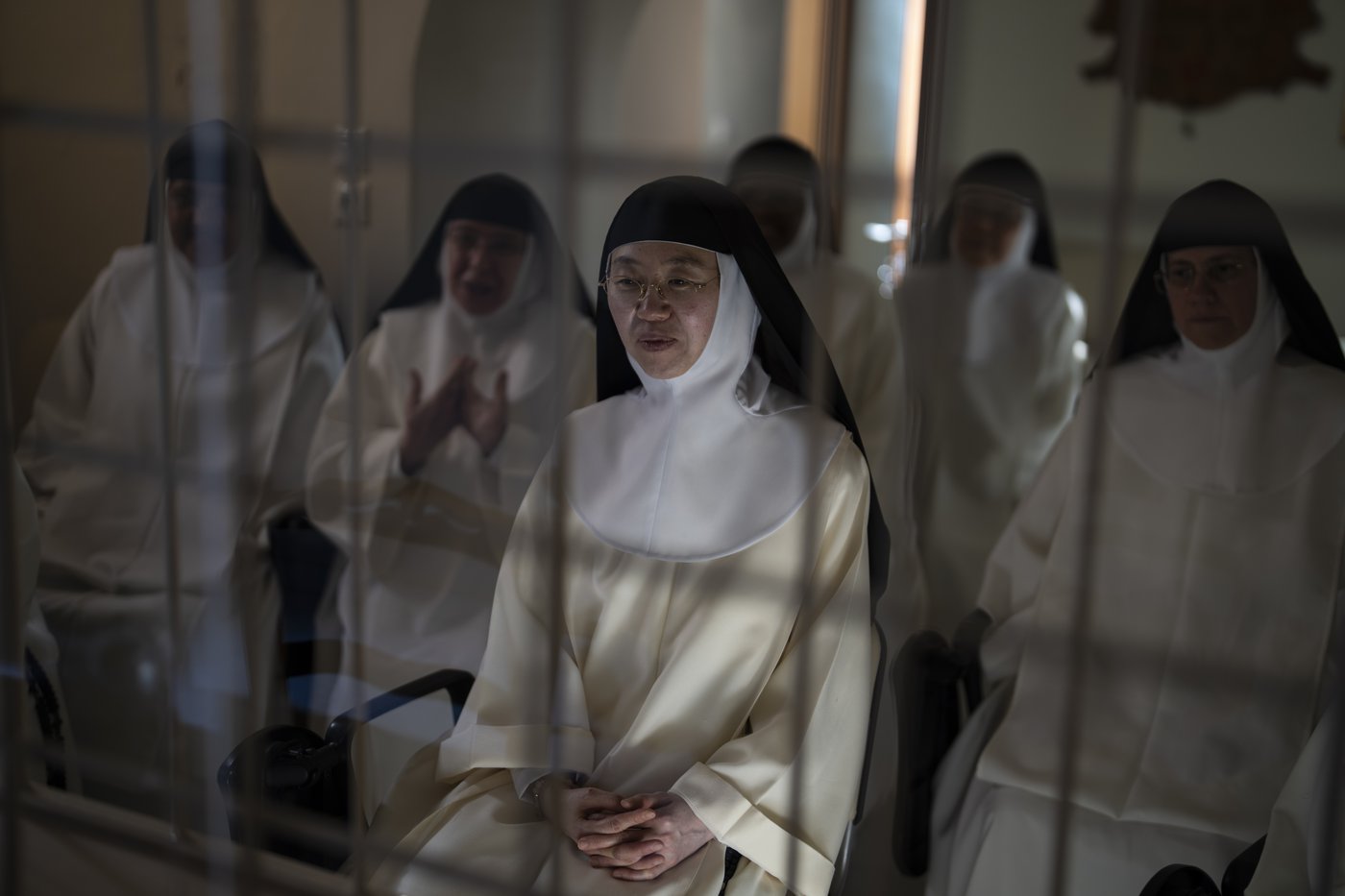 Nuns sitting in white robes with black head coverings sit in a couple rows together. They are seen through the openings of white iron bars