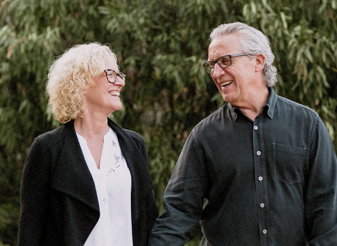 A man and a woman stand side-by-side, looking and smiling at each other. The woman has light skintone and blonde hair, the man has light skin tone and grey hair.
