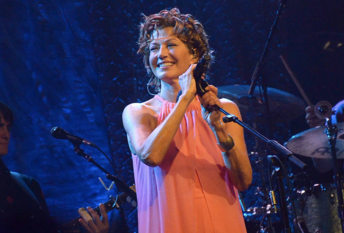 A woman with light skin tone grabs a microphone on a stand and smiles.