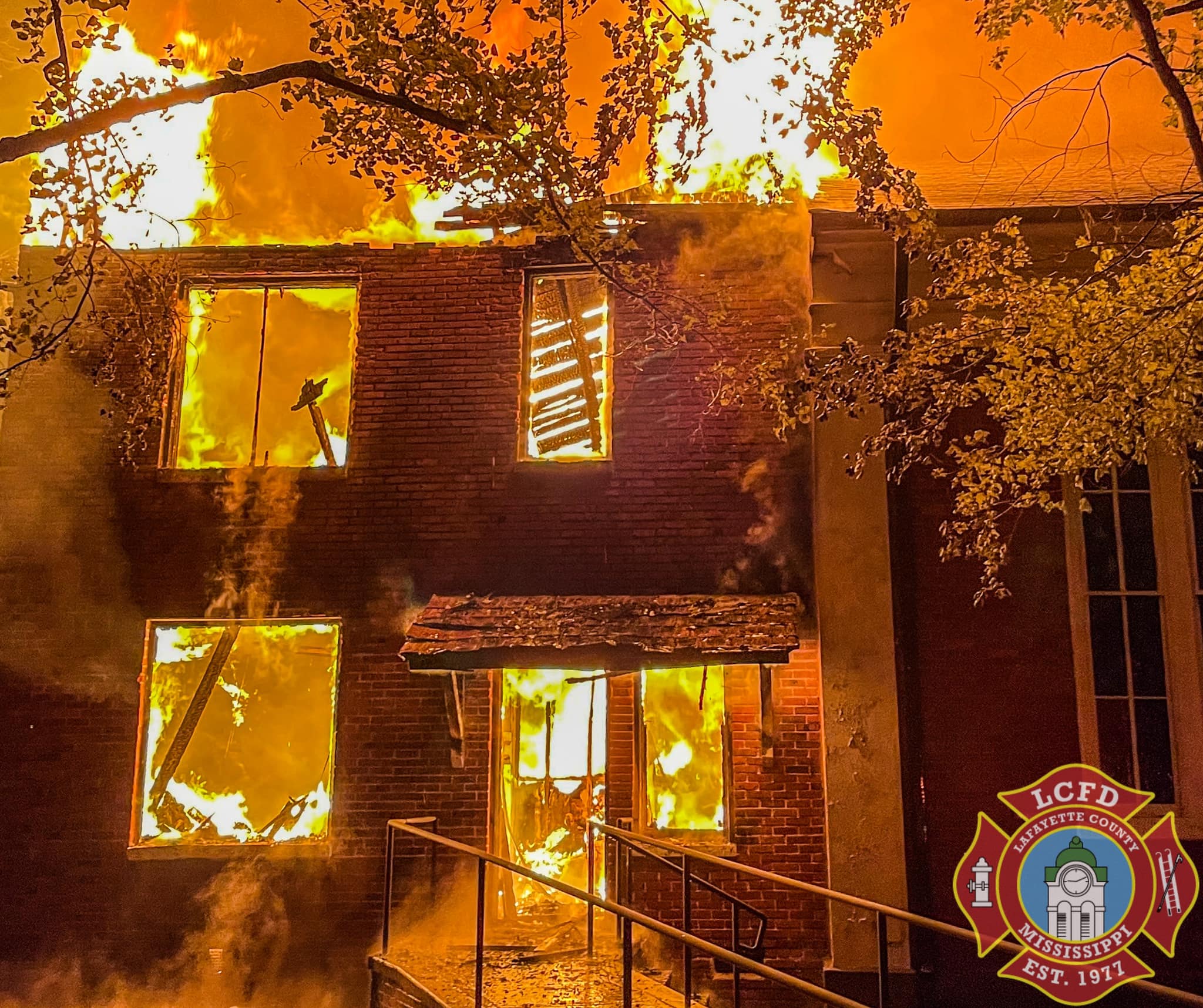 A close up of a brick building consumed by fire. large orange flames shoot up from the top. Windows and doorways are burned out with intense flames surrounding them.