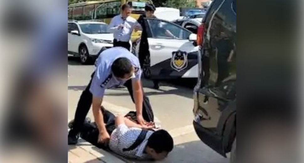 A man lays on the ground with a hand behind his back while an officer pushes down on his back while crouched above him. 2 officers are in the background opening a police car door.